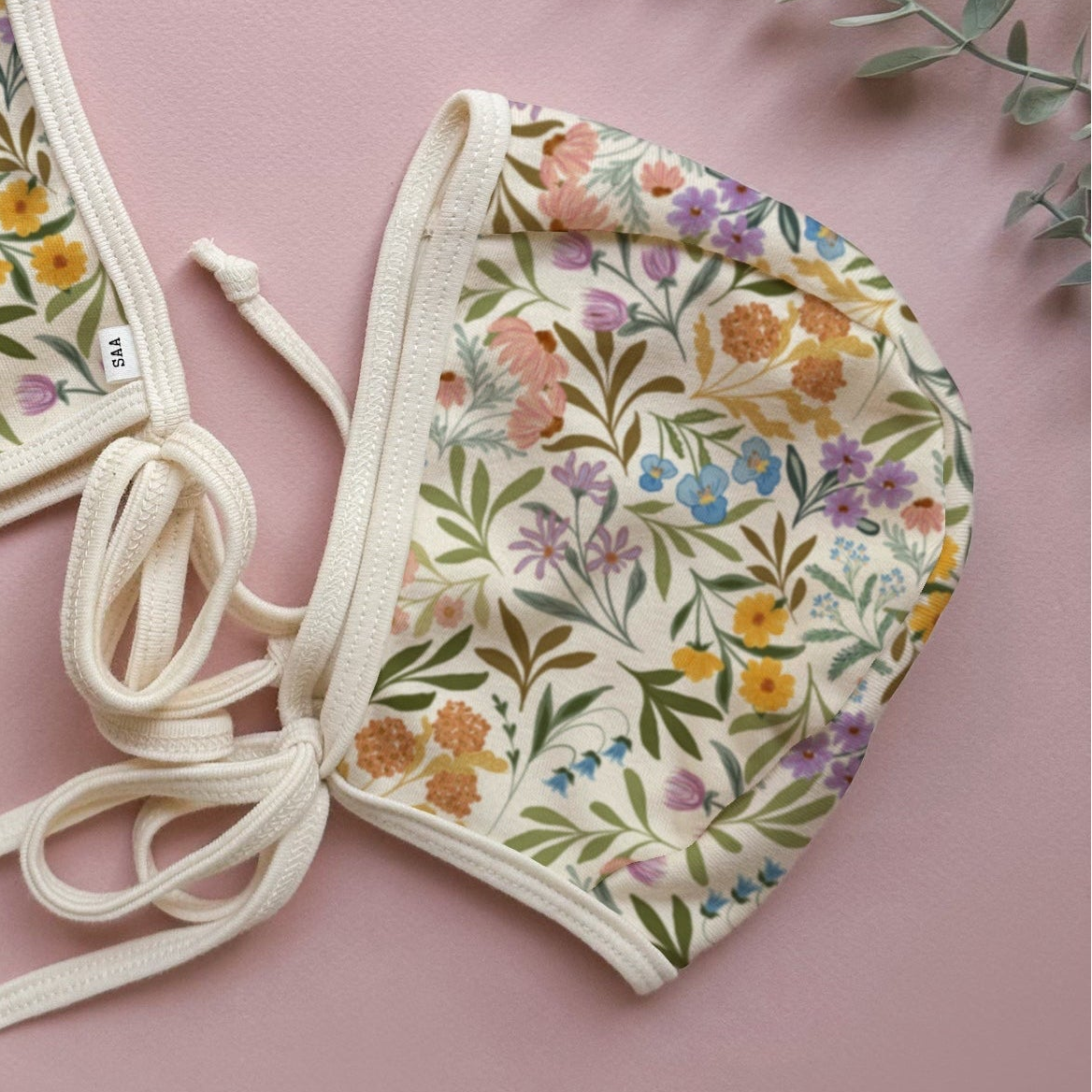 Floral-patterned baby bonnet on a pink background with green leaves.