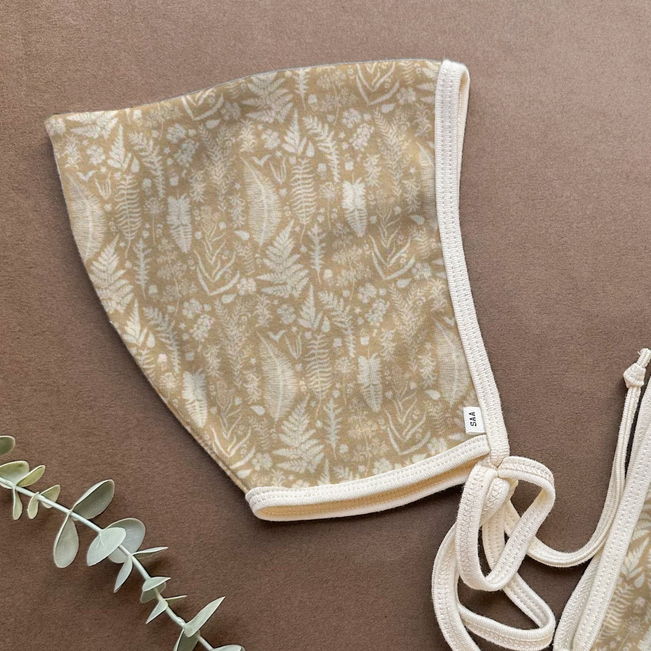 A pair of beige organic cotton baby bonnets with a leaf pattern, displayed on a brown background with green foliage.