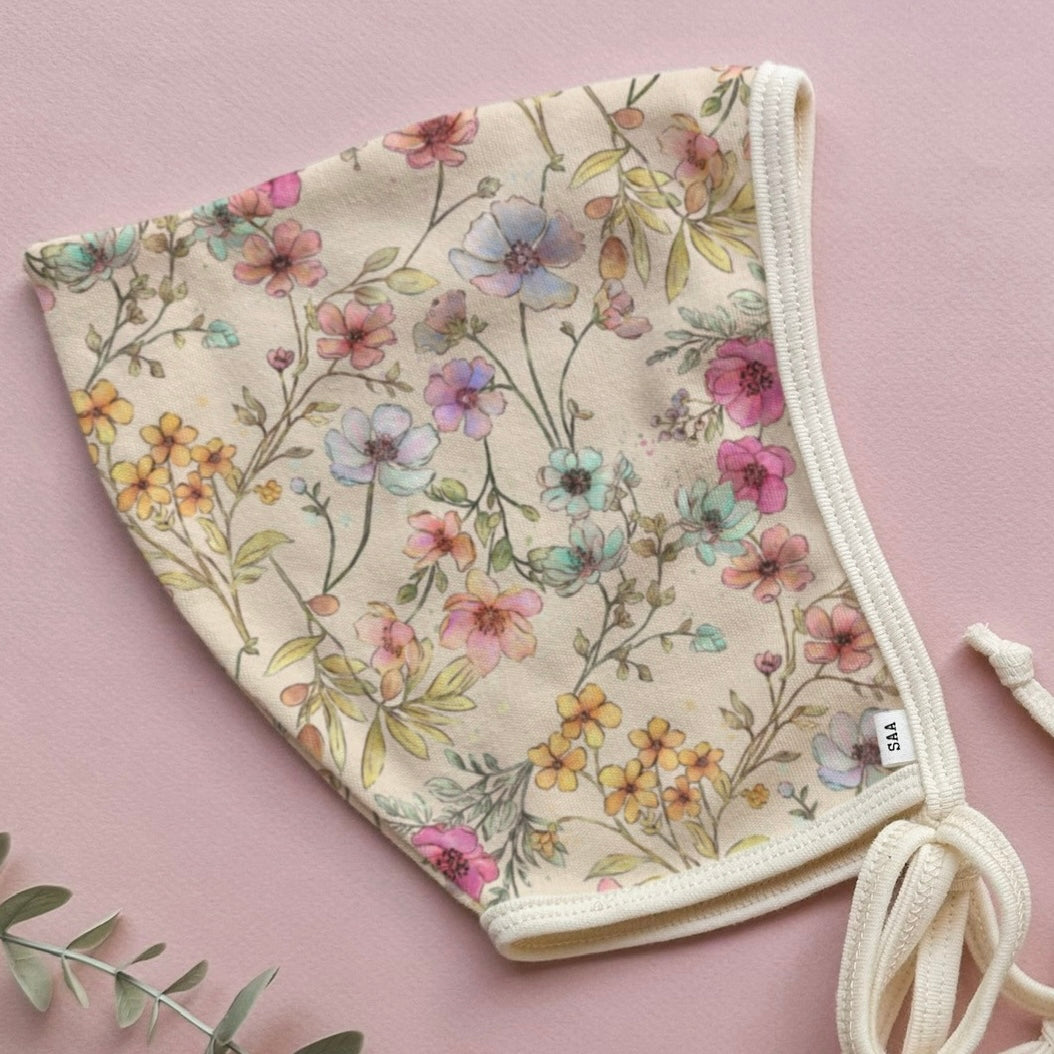 A floral patterned cotton baby bonnet on a pink background with some decorative greenery.