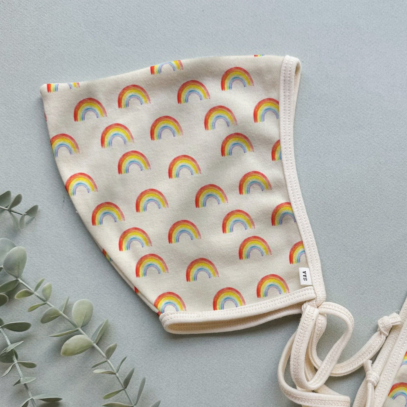 A pair of beige baby bonnets with a multicolored rainbow pattern on a fabric background, displayed on a flat surface with a few green leaves and a tie for adjustability.