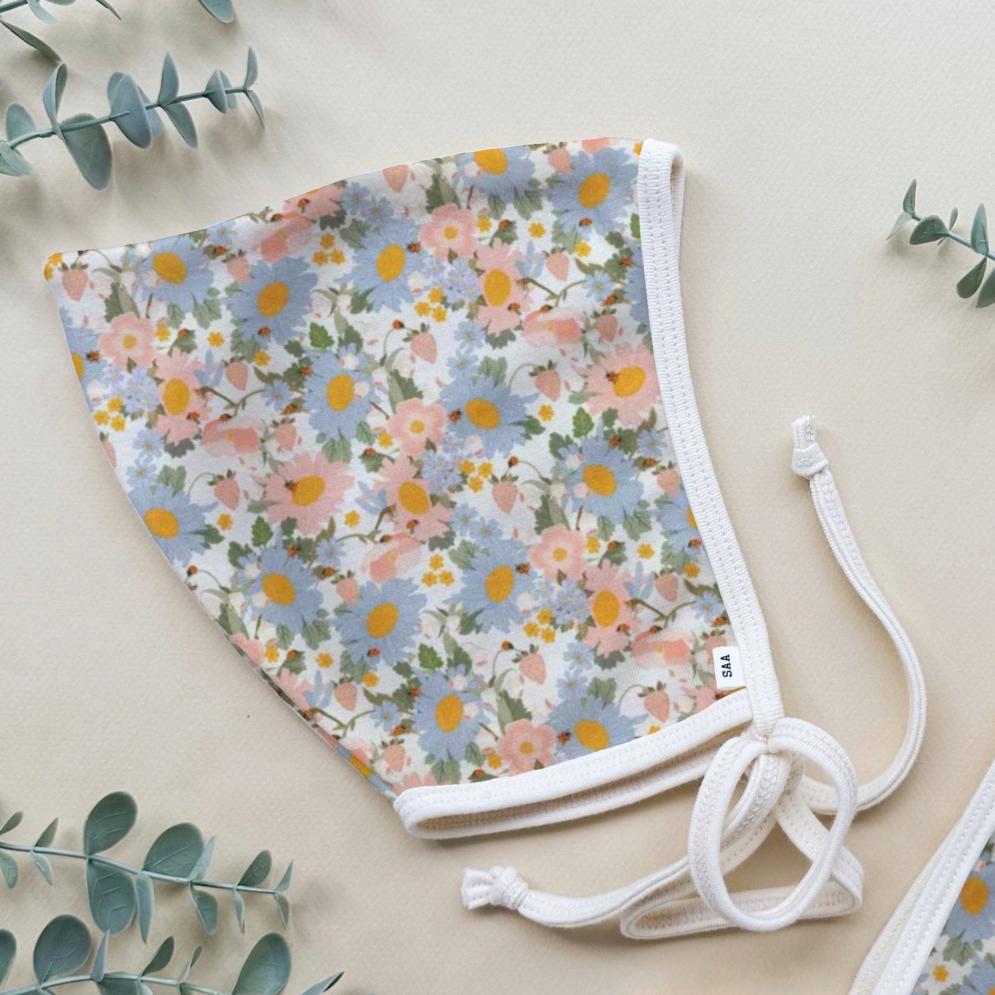 A pair of cotton baby bonnets with a floral pattern on a light background, featuring ties at the back for size adjustment, displayed on a flat surface with green foliage in the background.