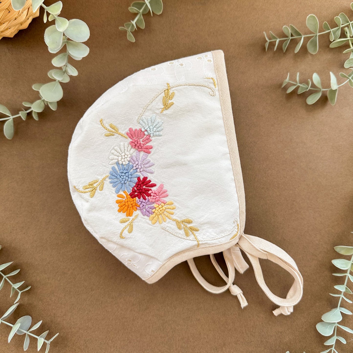 Embroidered baby bonnet with floral design on a brown background with eucalyptus leaves.