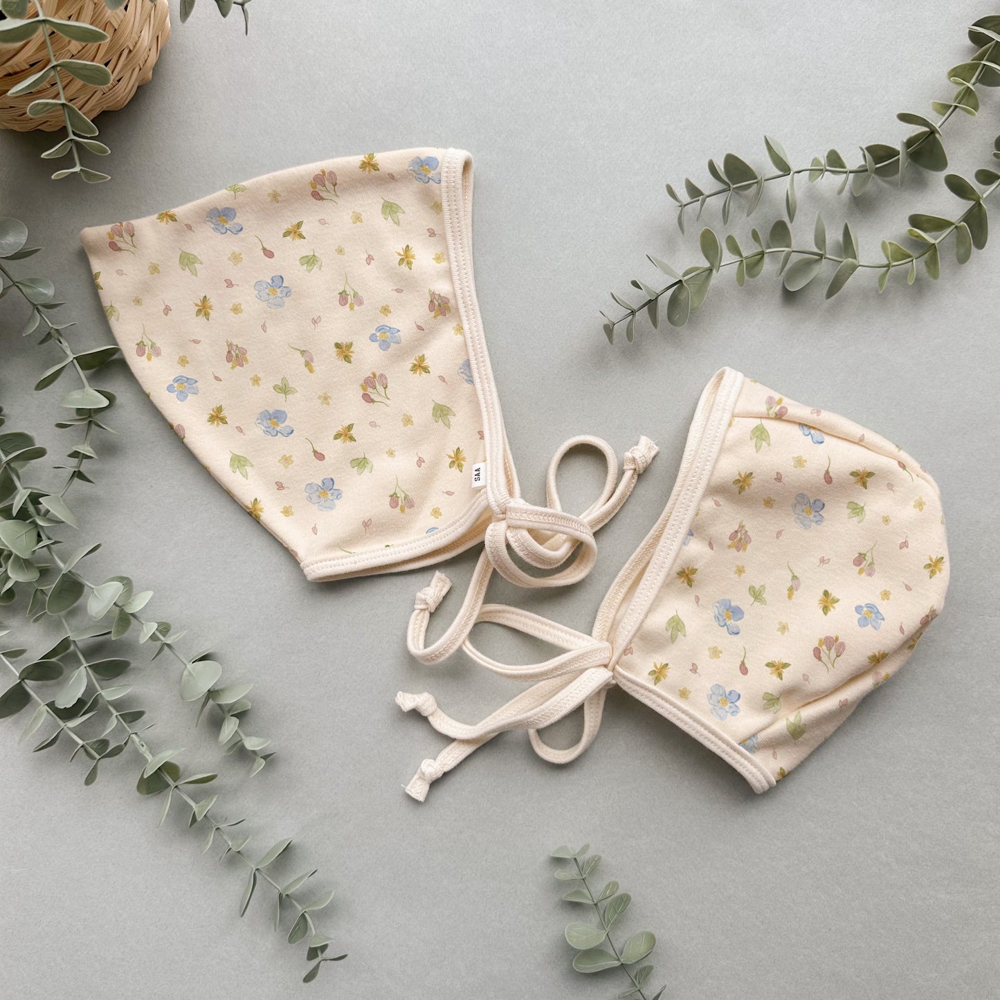 Two baby bonnets with floral pattern on a light gray background with green leaves.