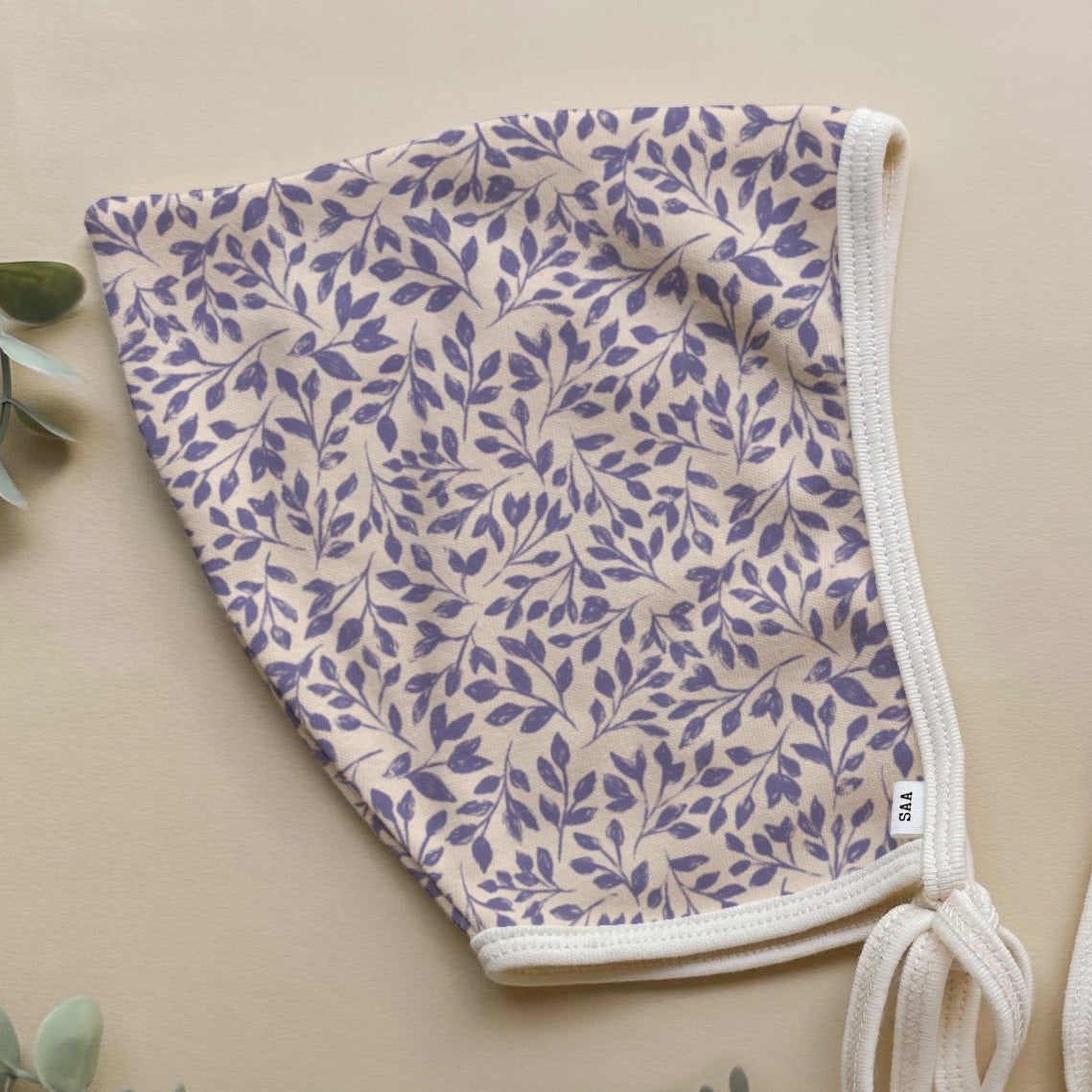 A floral patterned baby bonnet with a light background with some decorative greenary.