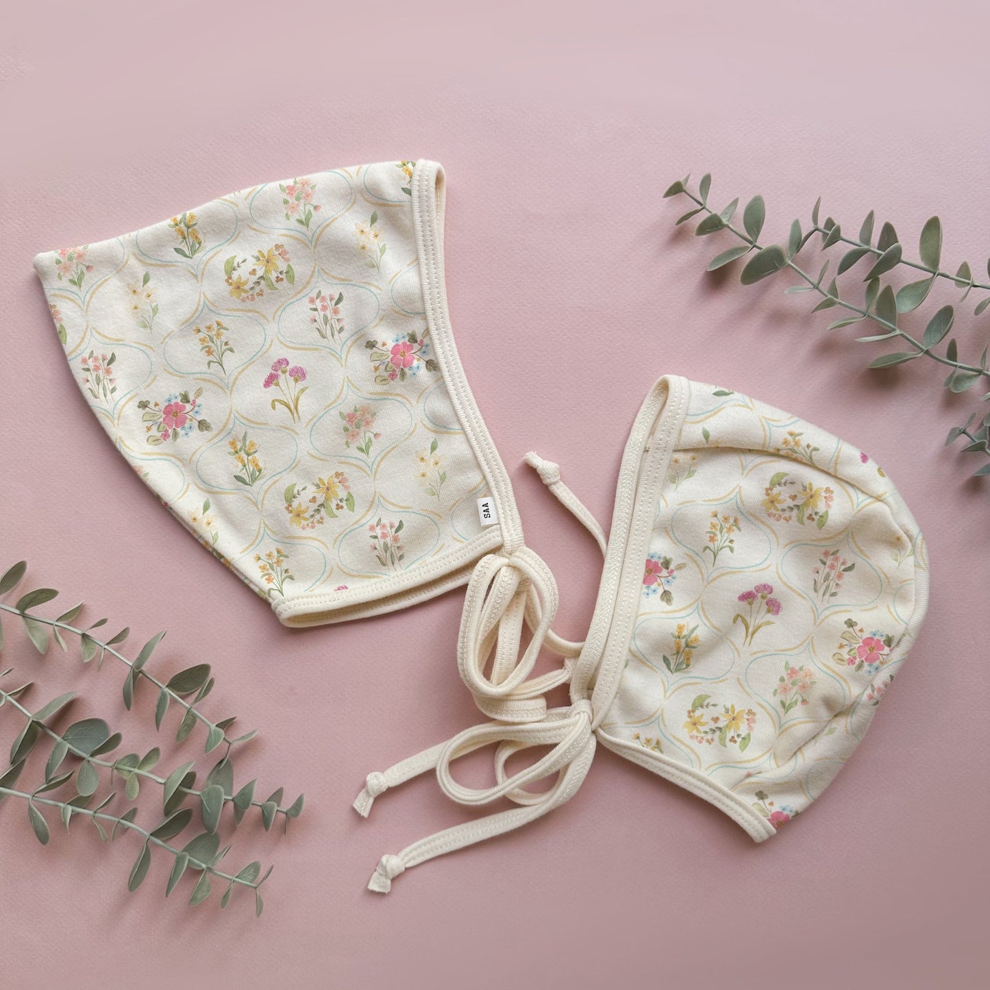 Two floral-patterned baby bonnets on a pink background with green leaves.