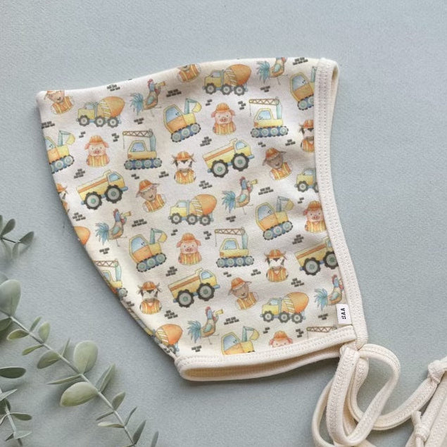 A pair of beige baby bonnets with a pattern of cartoonish vehicles on a gray background, displayed on a flat surface with some green foliage in the background.