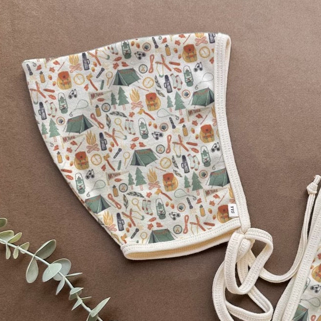 Two organic cotton baby bonnets with a floral and geometric pattern, displayed on a flat surface with some greenery in the background.