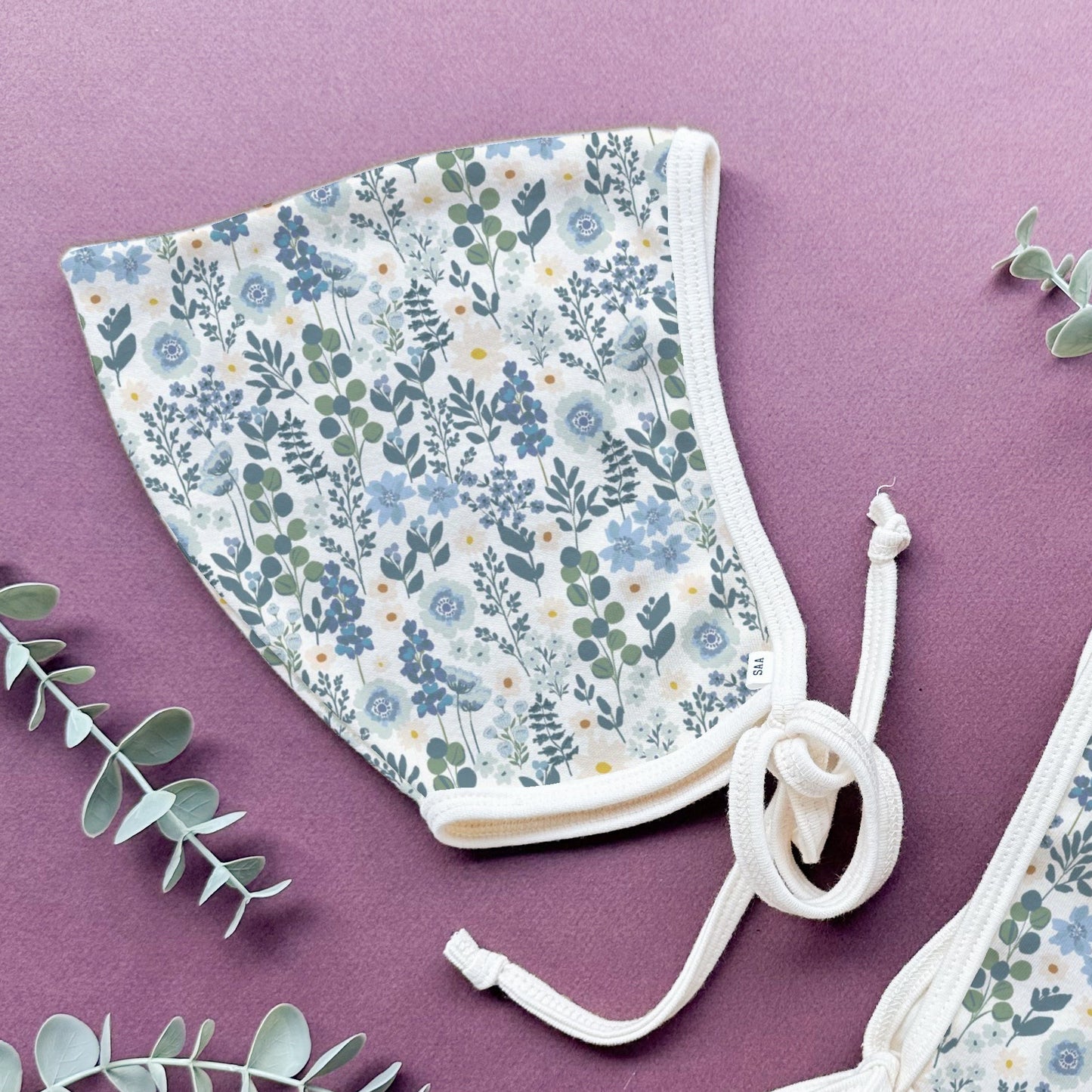 Two floral-patterned cotton baby bonnets on a purple background with eucalyptus leaves.