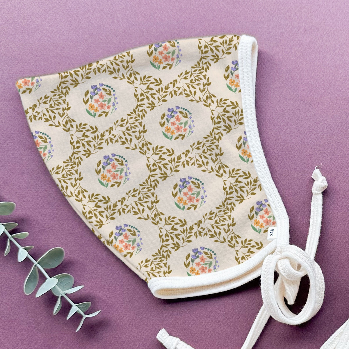 A floral patterned baby bonnet with a purple background with some decorstive greenary.