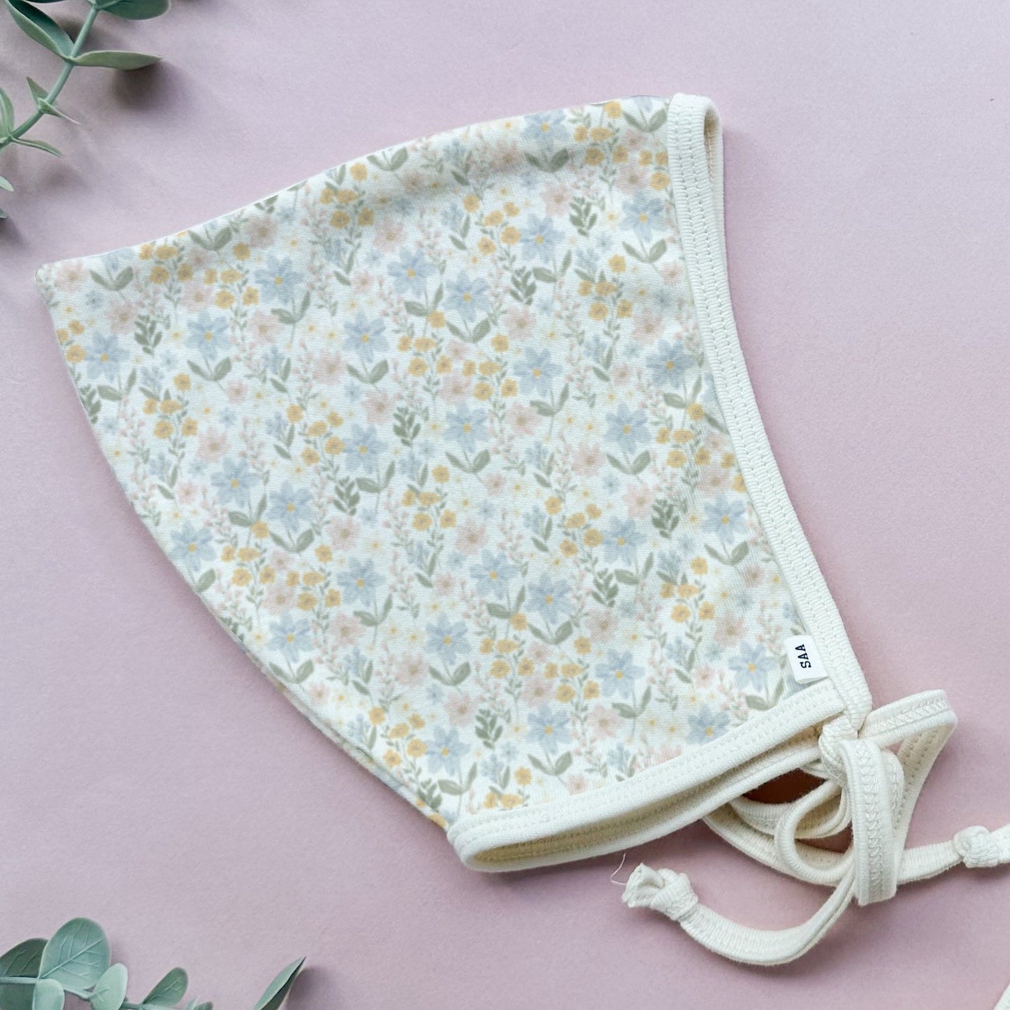 A floral patterned cotton baby bonnet on a pink background with some decorative greenery.