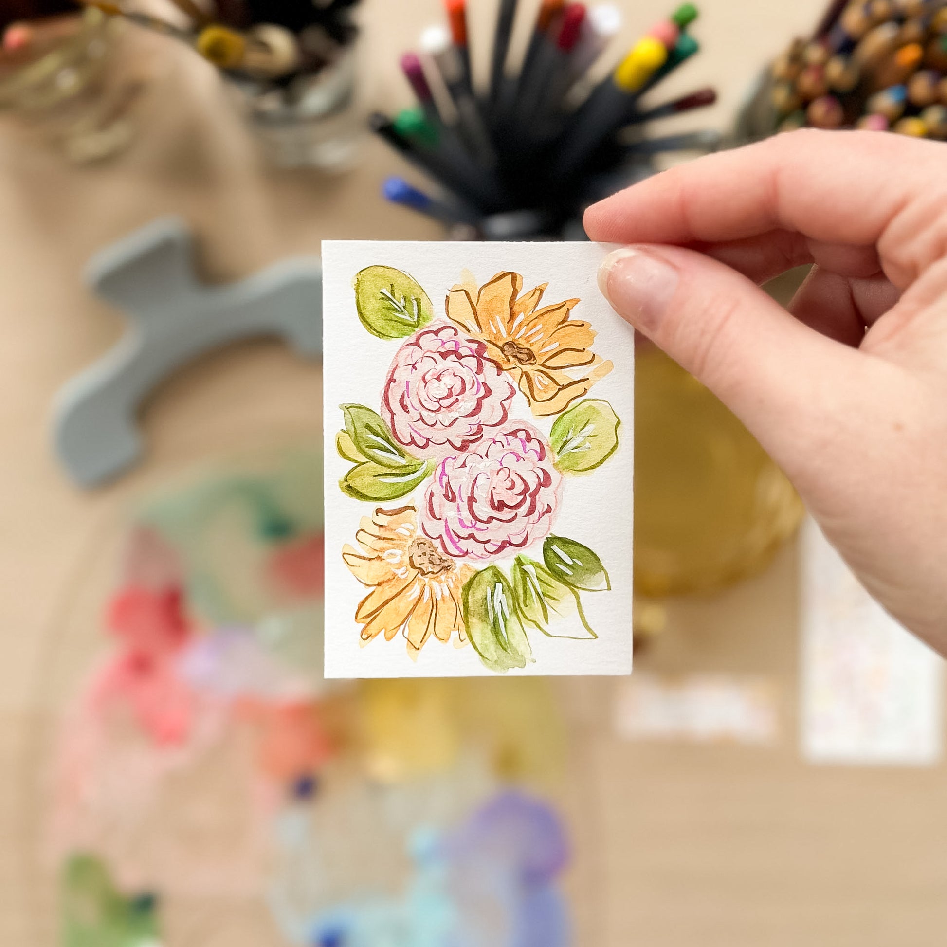 Hand holding a card with floral design in front of art supplies. ACEO original artwork by Carrie Keefe - Bloom by April