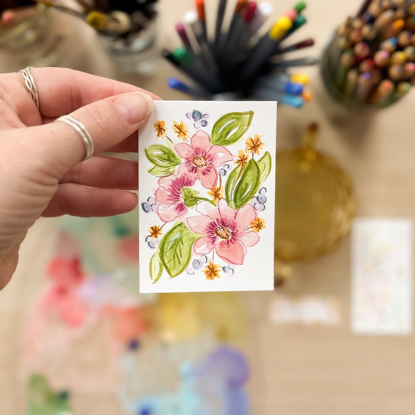 Hand holding a card with floral design in front of art supplies. ACEO original artwork by Carrie Keefe - Bloom by April