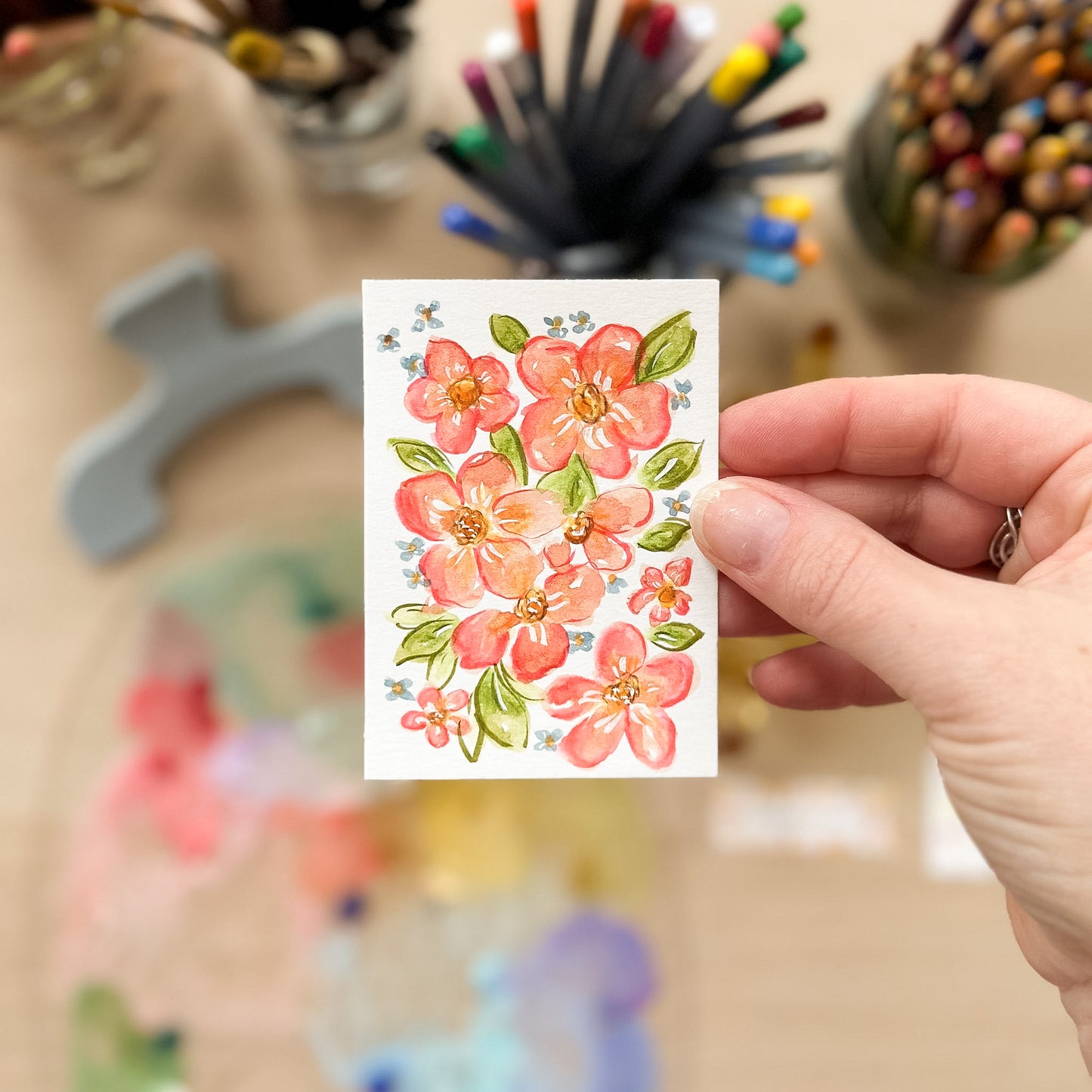 Hand holding a card with floral design in front of art supplies. ACEO original artwork by Carrie Keefe - Bloom by April