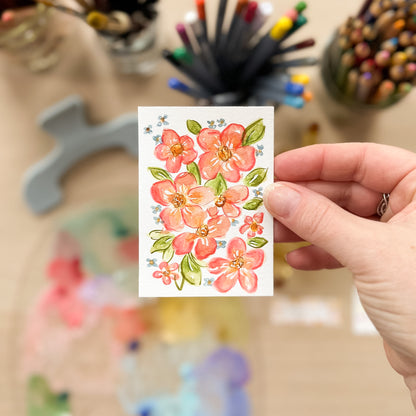 Hand holding a card with floral design in front of art supplies. ACEO original artwork by Carrie Keefe - Bloom by April