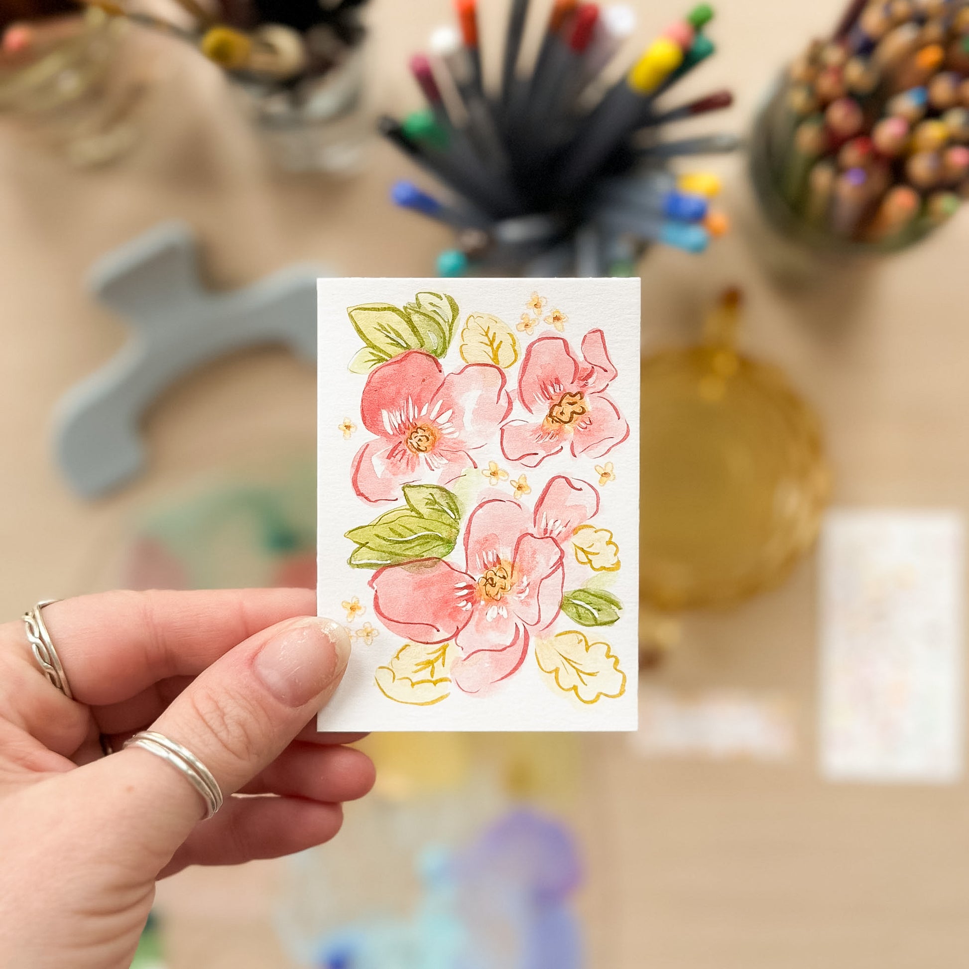 Hand holding a card with floral design in front of art supplies. ACEO original artwork by Carrie Keefe - Bloom by April