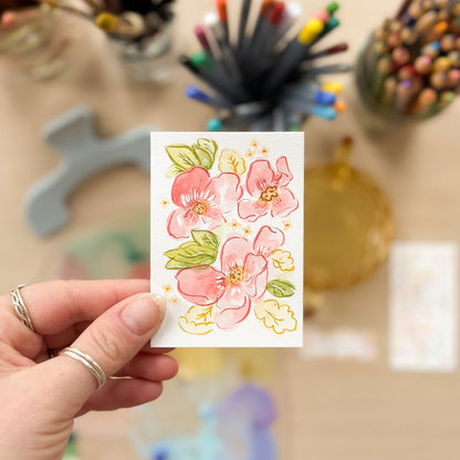Hand holding a card with floral design in front of art supplies. ACEO original artwork by Carrie Keefe - Bloom by April