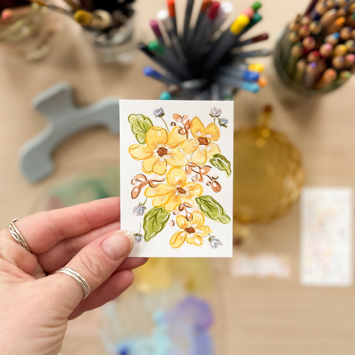 Hand holding a card with a floral design in front of art supplies on a blurred background. ACEO original artwork by Carrie Keefe - Bloom by April