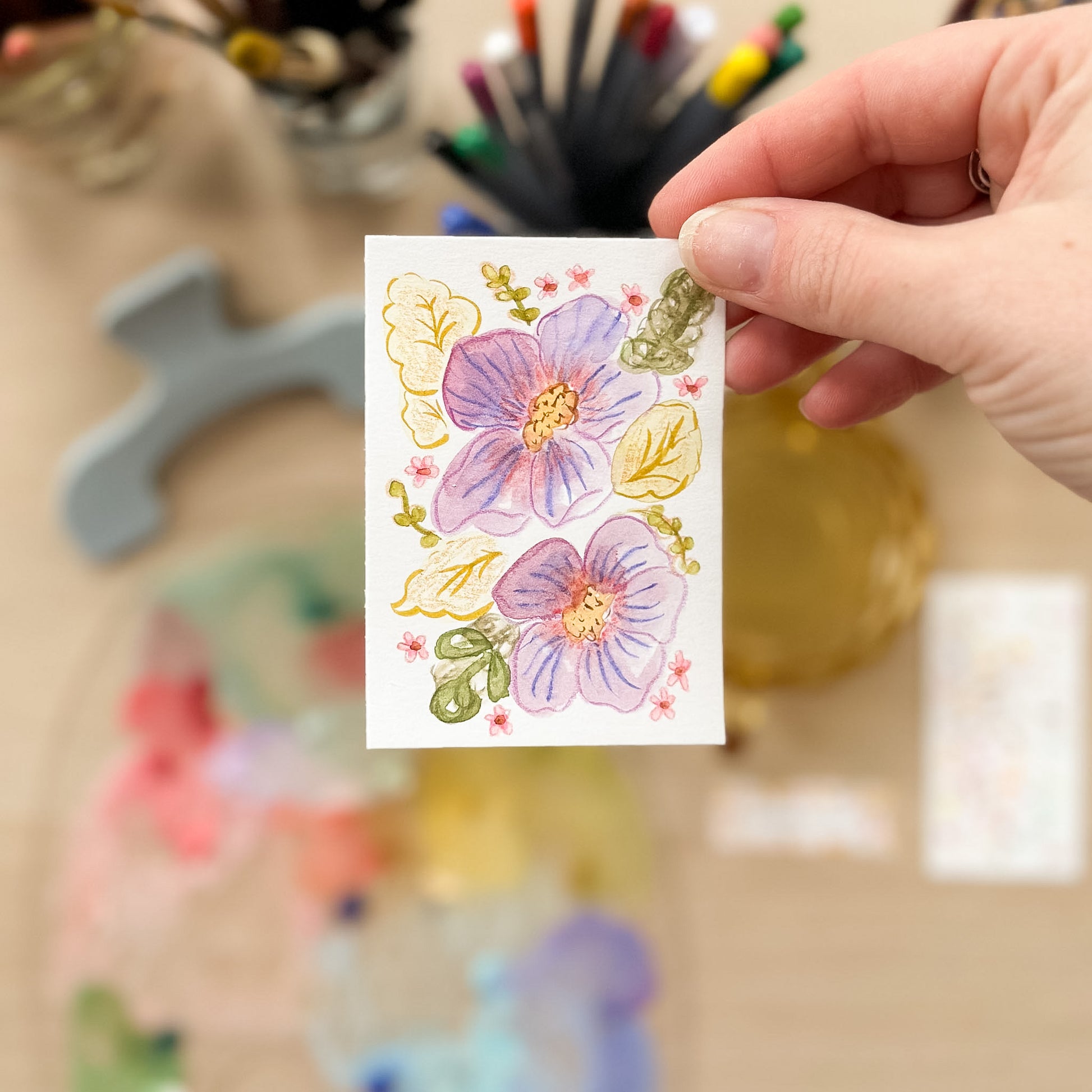 Hand holding a card with floral designs on a blurred background of art supplies. ACEO original artwork by Carrie Keefe - Bloom by April