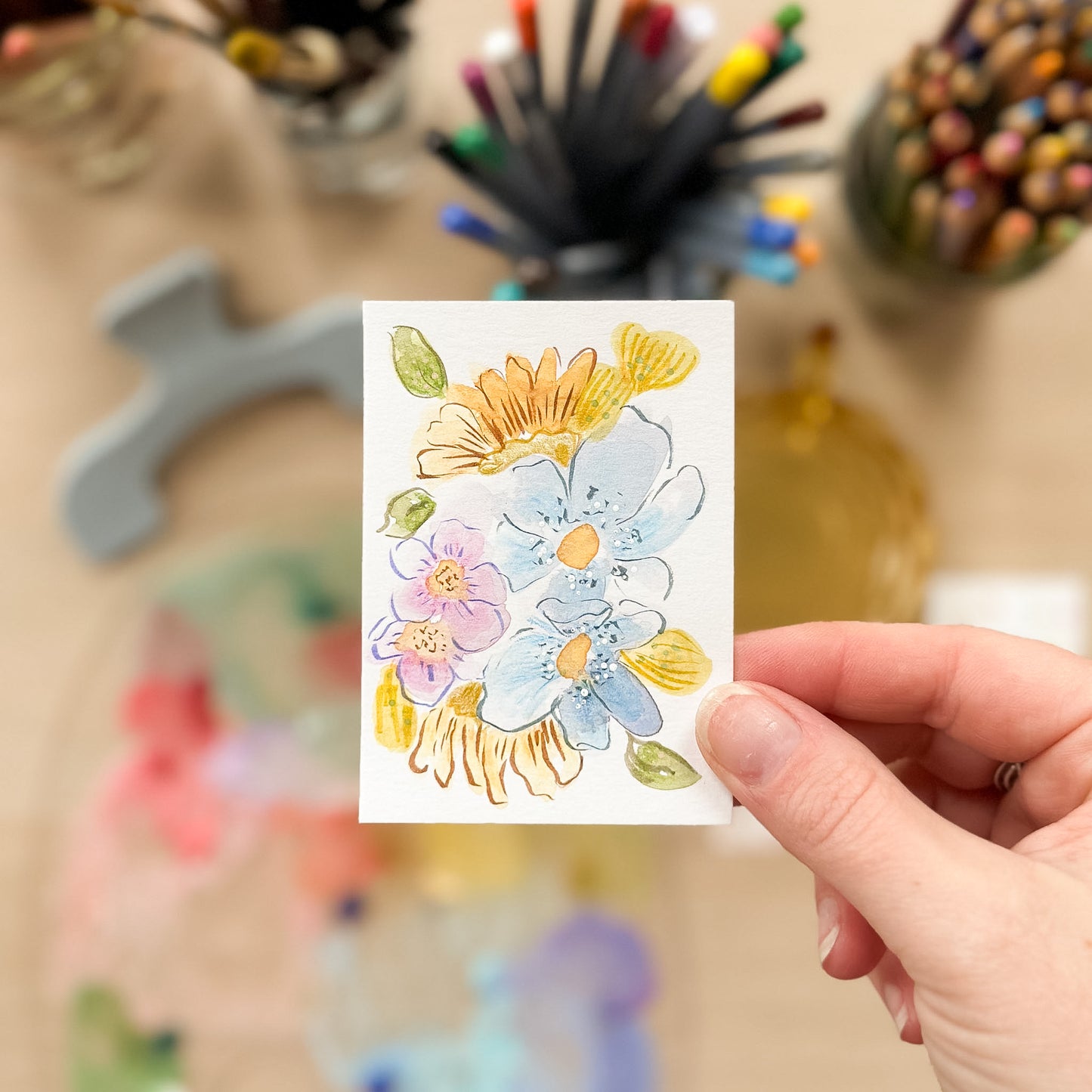 Hand holding a card with floral design in front of art supplies. ACEO original artwork by Carrie Keefe - Bloom by April