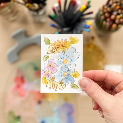Hand holding a card with floral design in front of art supplies. ACEO original artwork by Carrie Keefe - Bloom by April