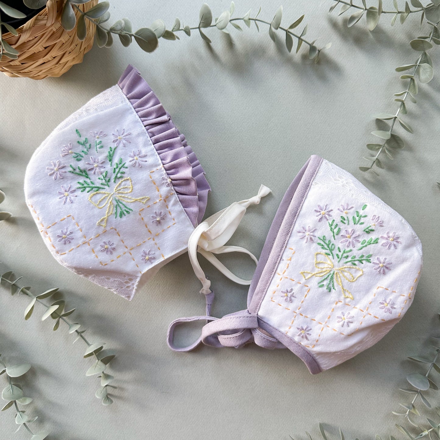 A pair of baby bonnets in a soft lavender and white fabric, decorated with embroidered flowers, bows, and a diamond lattice pattern. The bonnets are arranged on a light gray surface with decorative eucalyptus leaves and a wicker basket in the background.