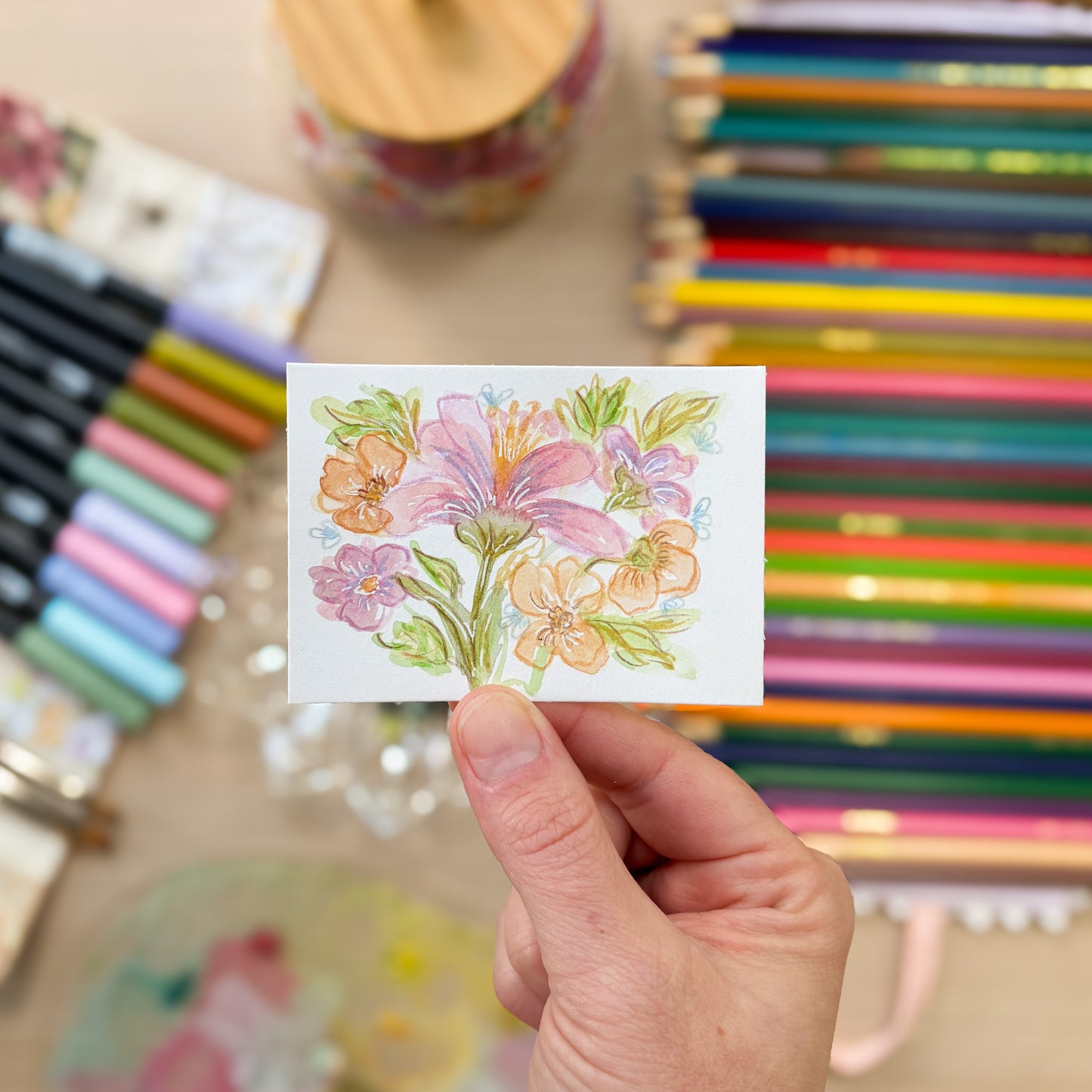 Hand holding a small card with a floral design in front of colored pencils and markers. ACEO original artwork by Carrie Keefe - Bloom by April