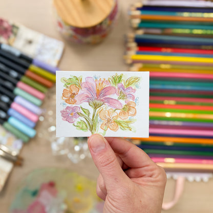 Hand holding a small card with a floral design in front of colored pencils and markers. ACEO original artwork by Carrie Keefe - Bloom by April