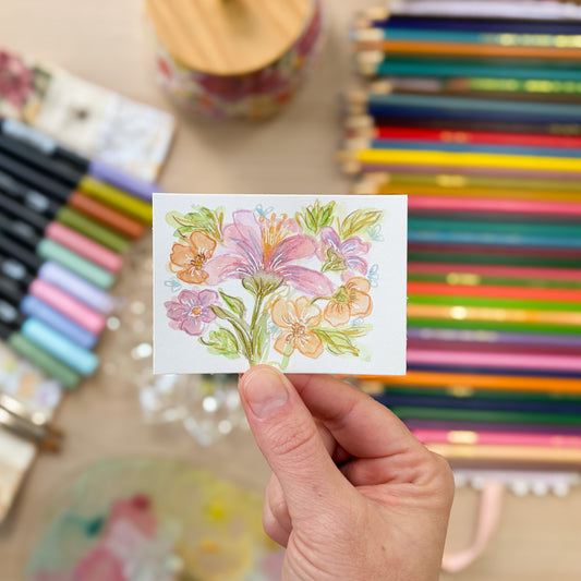 Hand holding a small card with a floral design in front of colored pencils and markers. ACEO original artwork by Carrie Keefe - Bloom by April