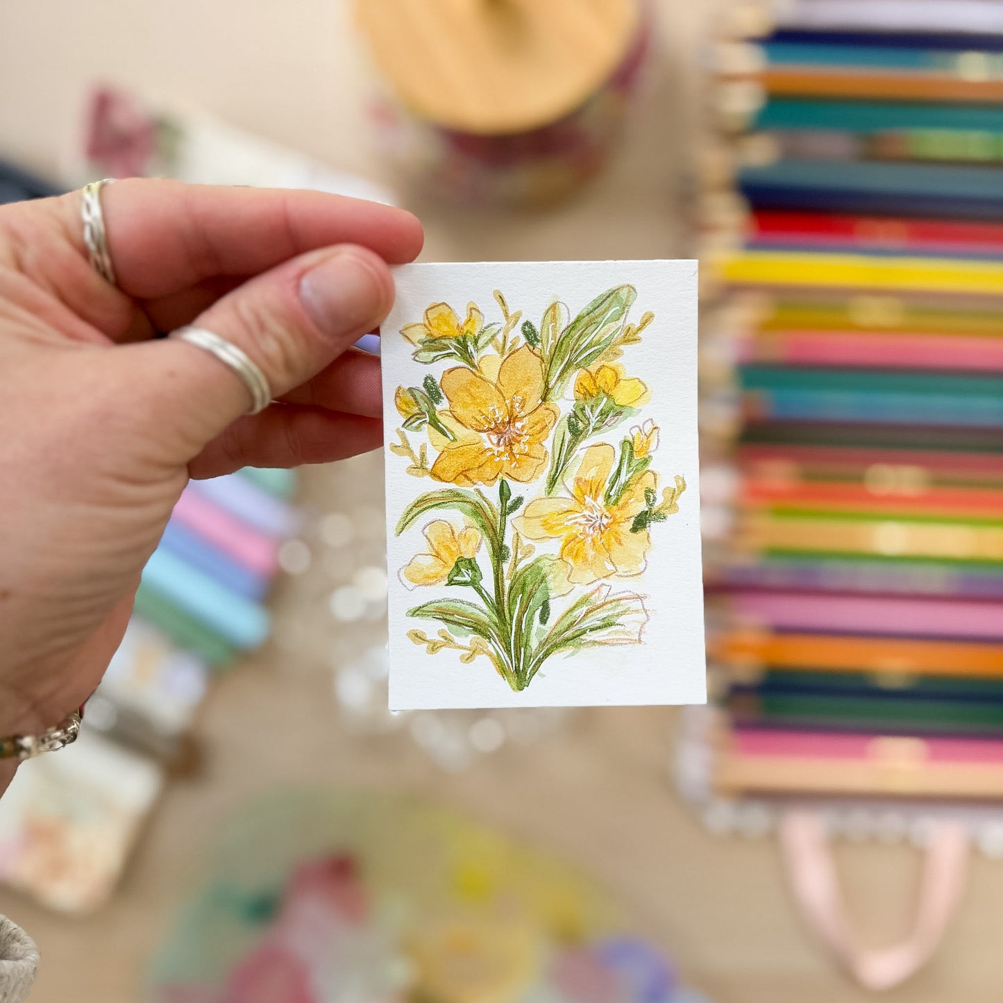 Hand holding a card with yellow floral design in front of colorful books. ACEO original artwork by Carrie Keefe - Bloom by April