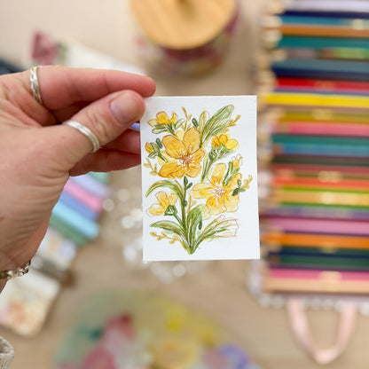 Hand holding a card with yellow floral design in front of colorful books. ACEO original artwork by Carrie Keefe - Bloom by April