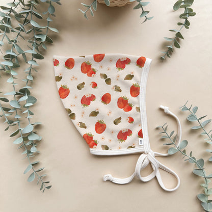 A strawberry patterned cotton baby bonnet on a beige background with some decorative greenery.