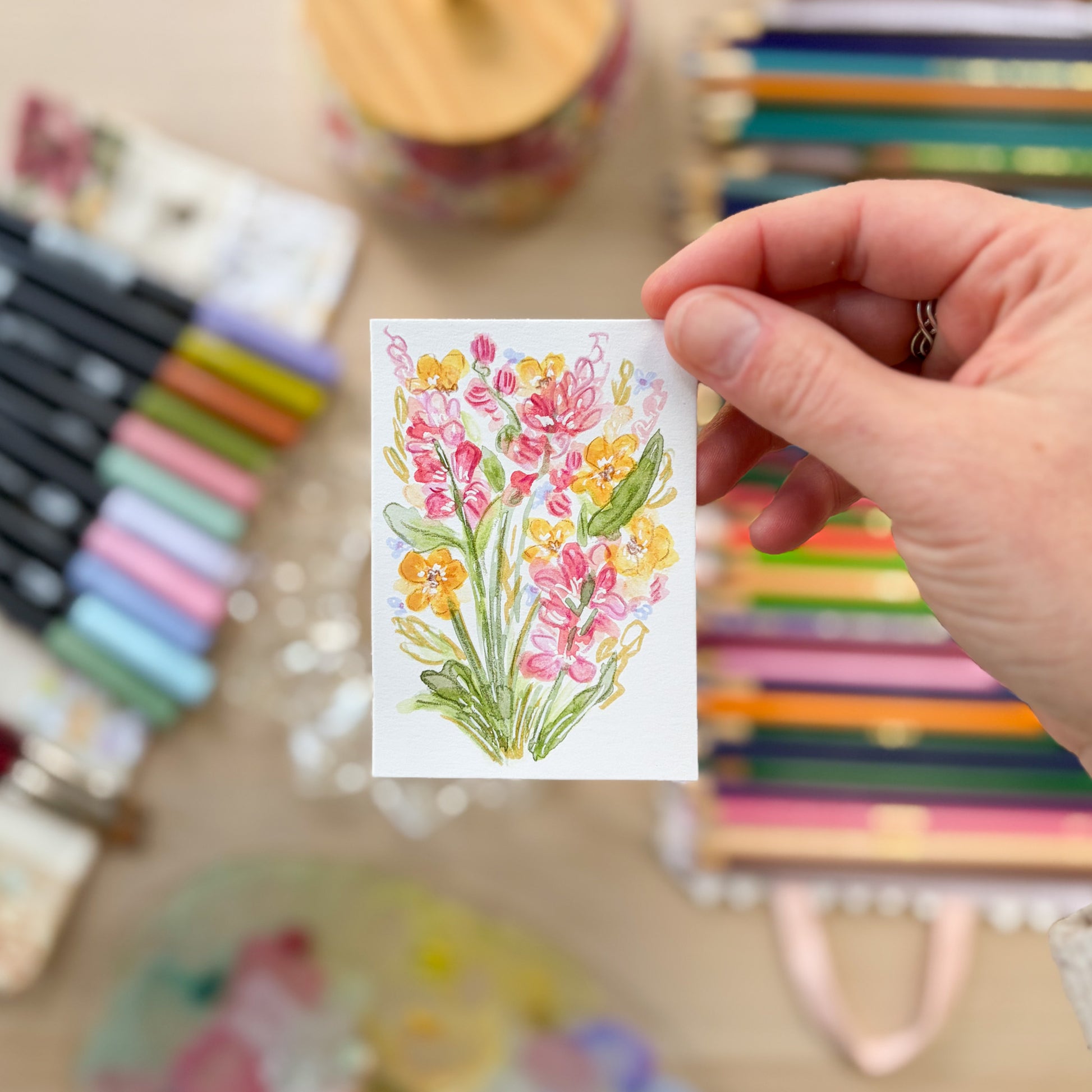 Hand holding a card with floral design surrounded by stationery items. ACEO original artwork by Carrie Keefe - Bloom by April