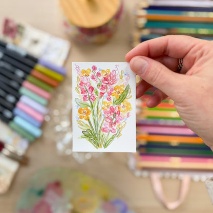 Hand holding a card with floral design surrounded by stationery items. ACEO original artwork by Carrie Keefe - Bloom by April