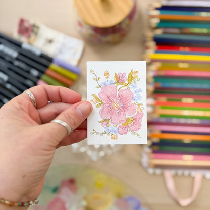 Hand holding a card with floral design in front of colorful stationery items. ACEO original artwork by Carrie Keefe - Bloom by April
