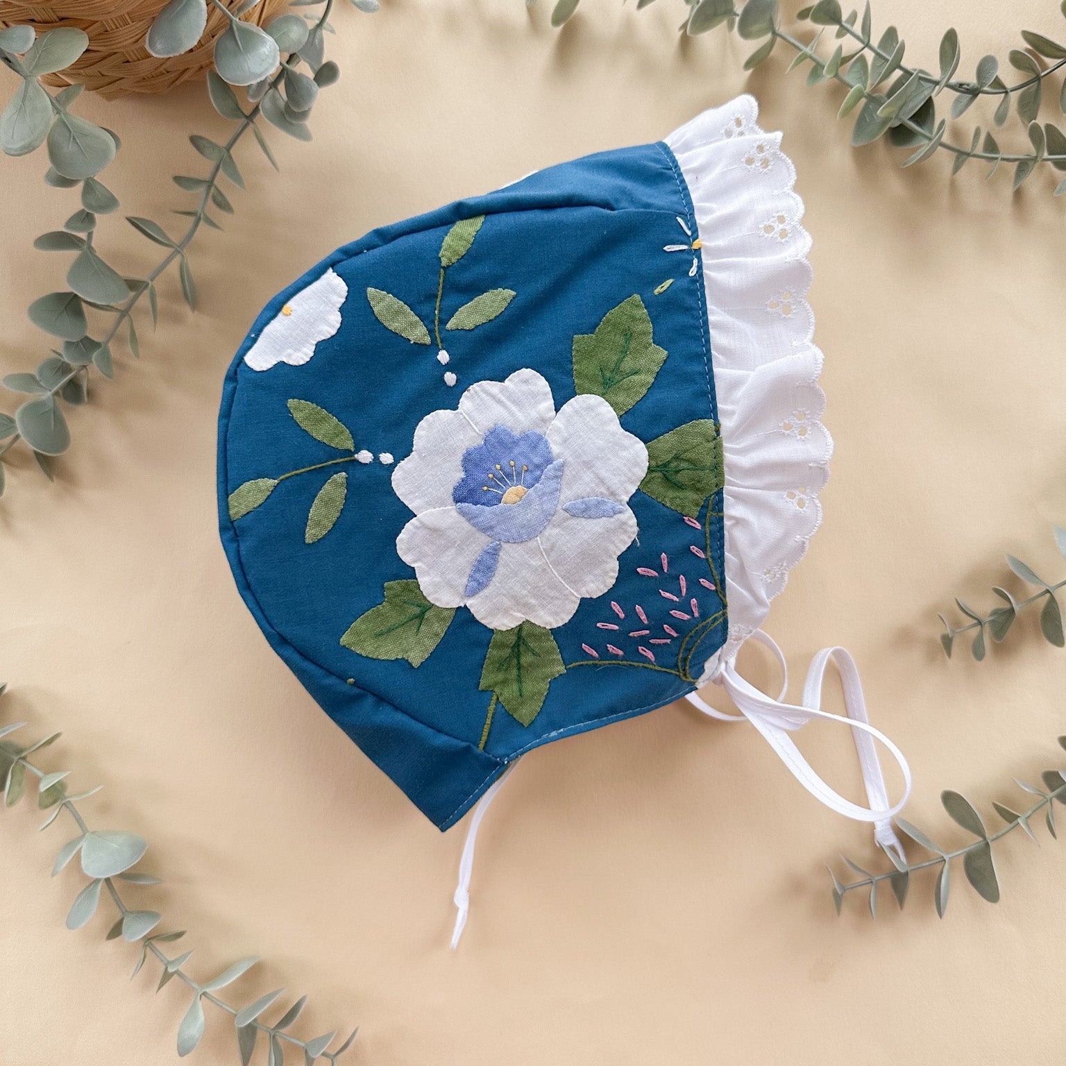 Blue floral baby bonnet on a beige background with green leaves.