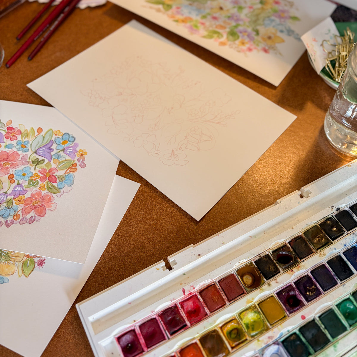 A watercolor paint palette with multiple colors, several sketch and painted paper sheets on a wooden table, and paintbrushes nearby.