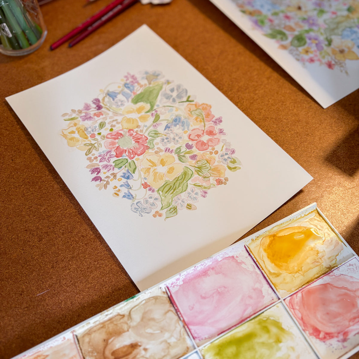A watercolor painting of a colorful floral bouquet on white paper, displayed on a brown cork desk alongside a watercolor palette with pink, yellow, green, and blue paints.