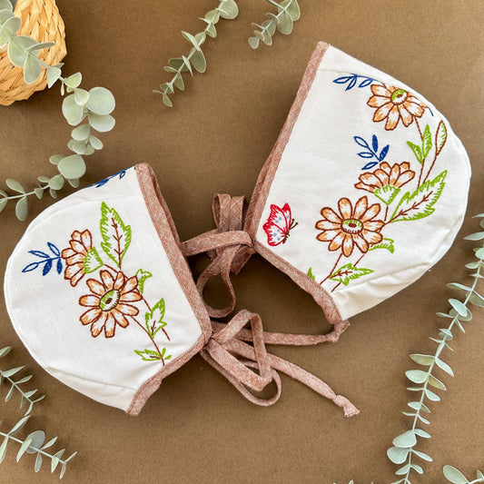 Two embroidered baby bonnets on a brown background with eucalyptus leaves.