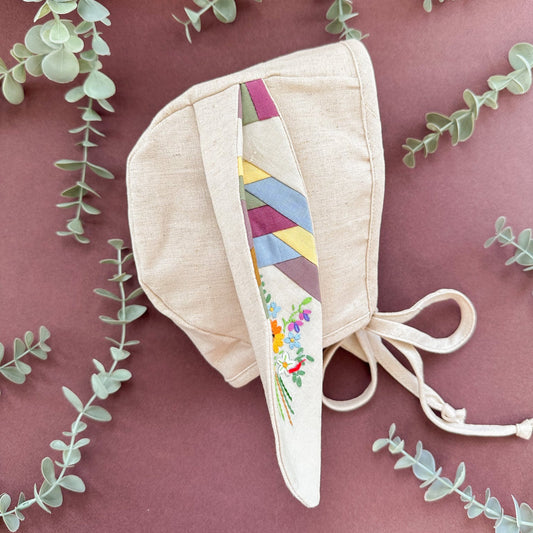 Beige baby bonnet with colorful ribbon on a pink background with eucalyptus leaves.