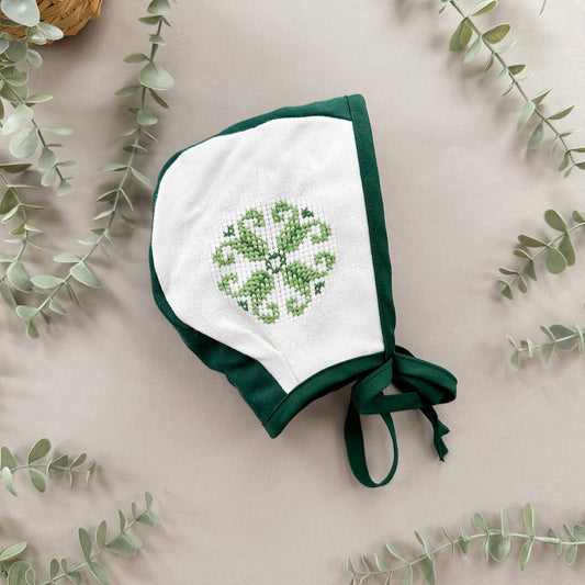 Green baby bonnet with shamrock design on a beige background with eucalyptus leaves.