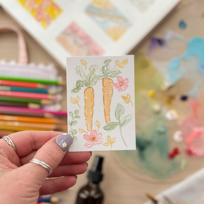 Hand holding a card with carrot illustrations and flowers against a colorful blurred background. ACEO original artwork by Carrie Keefe - Bloom by April