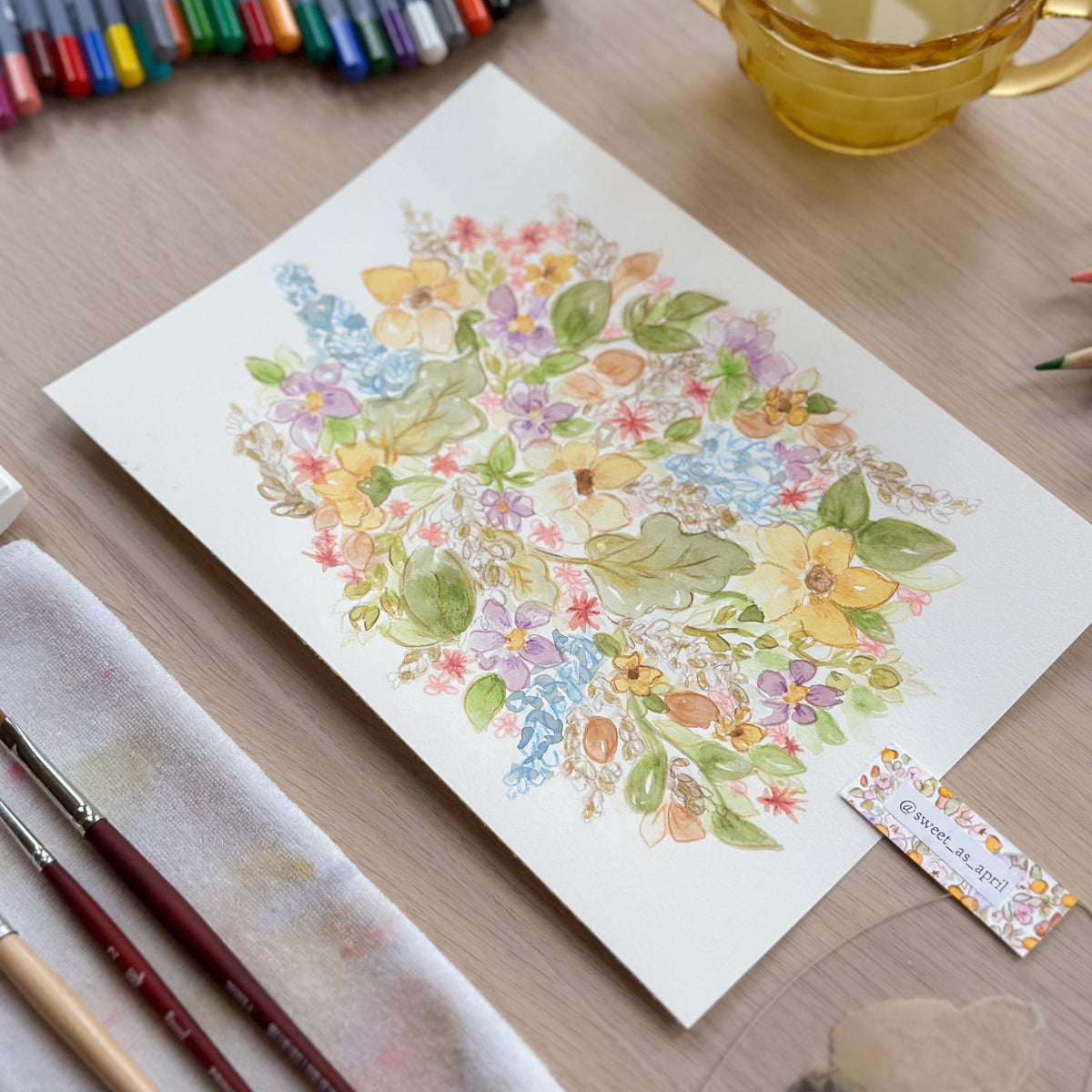 A watercolor painting of a colorful floral bouquet on a white sheet of paper, styled on a wooden desk with art supplies including colored pencils, paintbrushes, a yellow glass cup, and a palette.