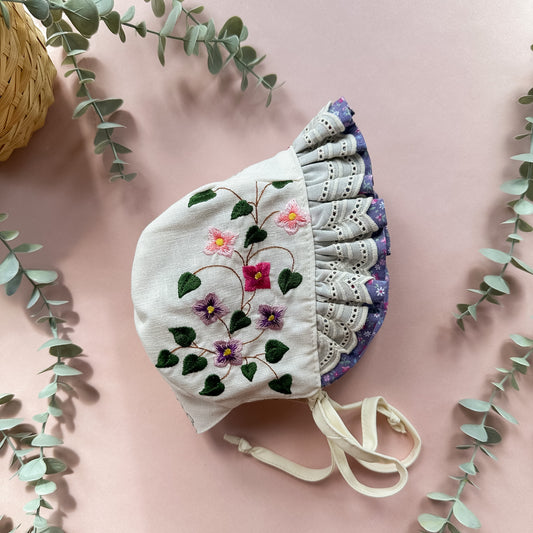Embroidered bonnet with floral design on a pink background with green leaves.