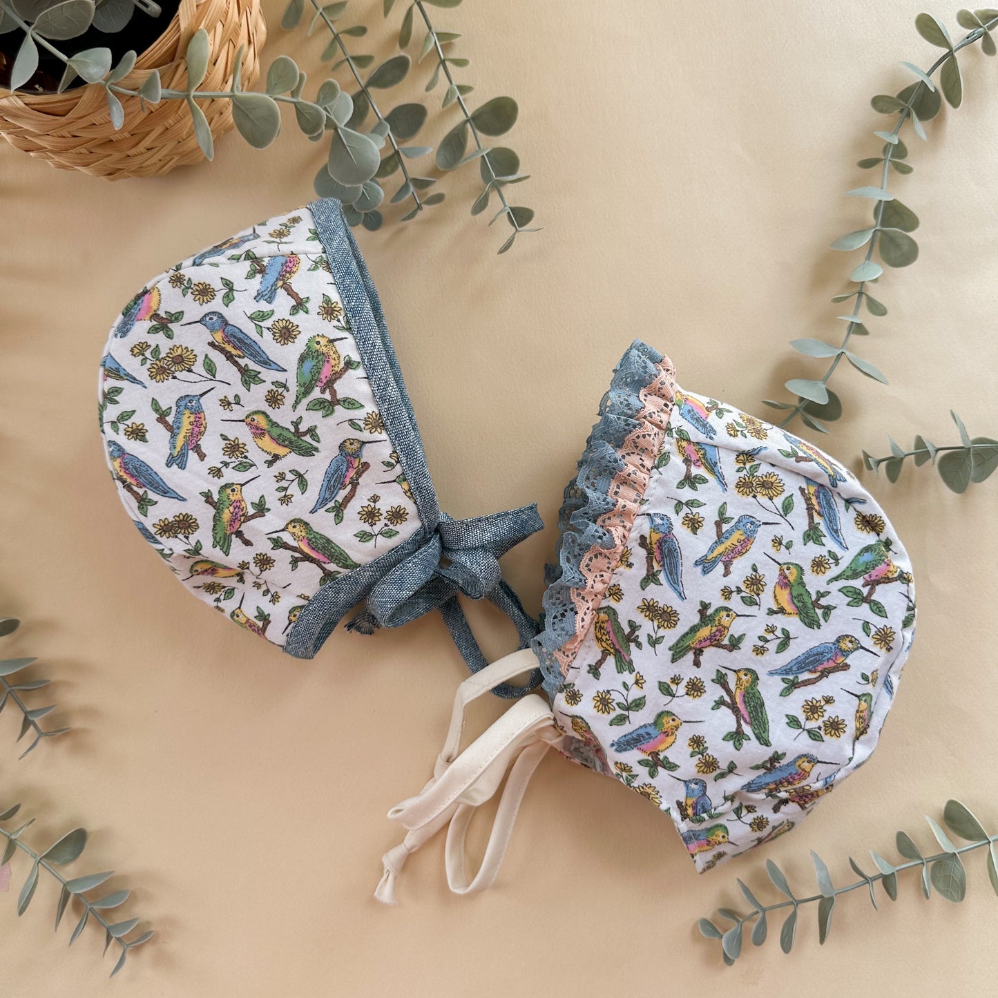 Two baby bonnets with bird pattern on a beige background with green leaves.