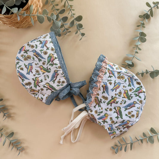 Two baby bonnets with bird pattern on a beige background with green leaves.