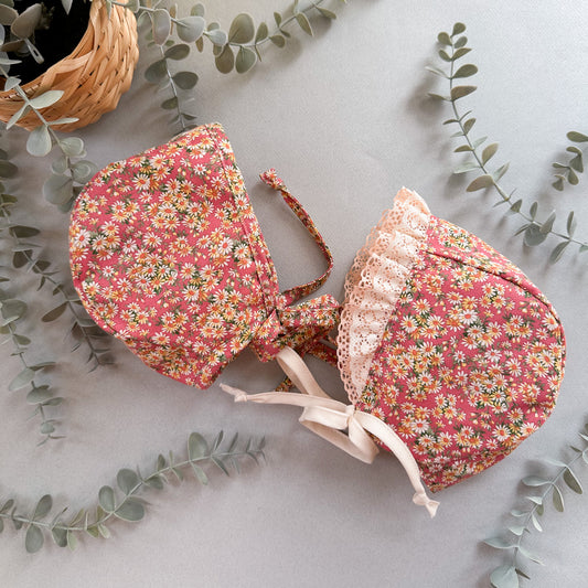 Two floral baby bonnets on a light surface with eucalyptus leaves.