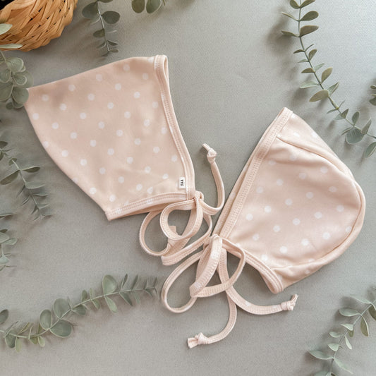 Two neutral polka dot baby bonnets on a light gray background with green leaves.