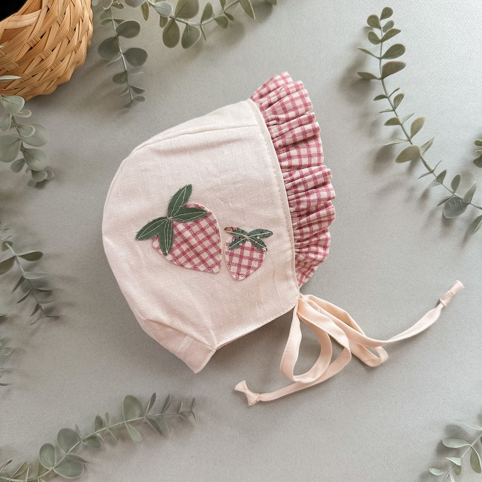 Baby bonnet with strawberry design on a light gray background with green leaves.