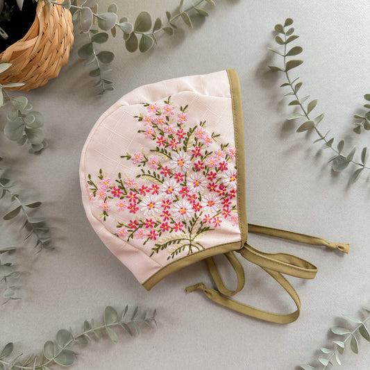 Floral embroidered baby bonnet on a light background with eucalyptus leaves.