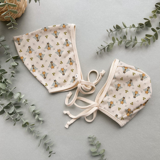 Baby bonnet with bee pattern on a light gray background with eucalyptus leaves.